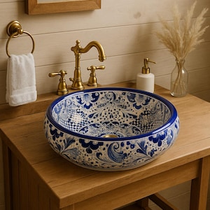 Puede incluir: Un lavabo redondo de cerámica azul y blanco con un patrón floral decorativo. El lavabo está colocado sobre un mueble de baño de madera con un grifo dorado y una toalla blanca. El lavabo mide aproximadamente 40 cm de diámetro.