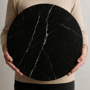 Puede incluir: Una mesa redonda de mármol negro con vetas blancas. La mesa es sostenida por una persona. El mármol tiene un acabado pulido y un aspecto de piedra natural. Adecuada para una mesa de comedor o de centro, de unos 76 cm de diámetro.