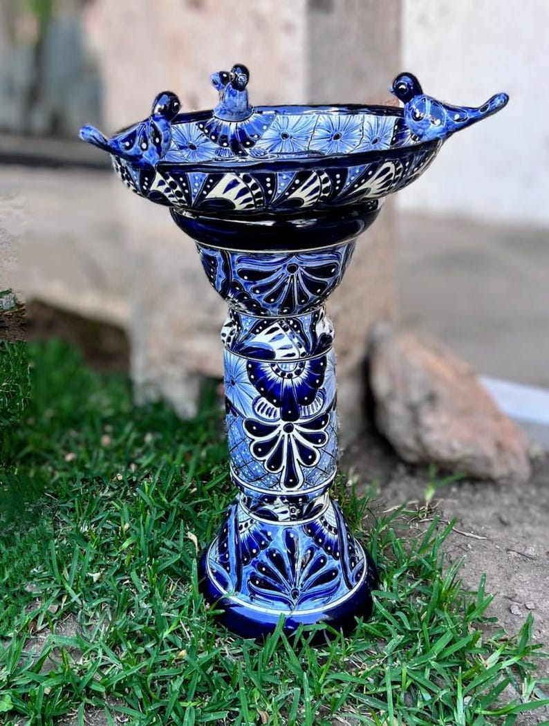 Pode incluir: Banho de p&aacute;ssaros de cer&acirc;mica azul e branco com tr&ecirc;s p&aacute;ssaros empoleirados na borda. O banho de p&aacute;ssaros est&aacute; em um suporte alto e decorativo com um padr&atilde;o floral azul e branco.