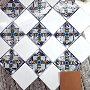 Puede incluir: Una colección de baldosas cerámicas cuadradas con un diseño floral decorativo en azul, amarillo y rojo sobre un fondo blanco. Las baldosas están dispuestas en un patrón de cuadrícula, con algunas baldosas que presentan el diseño completo y otras en blanco liso. También hay una sola baldosa marrón.