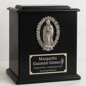 Puede incluir: Urna conmemorativa negra con un emblema religioso plateado y una placa con el nombre. La placa dice "Margarita Guzmán Gómez" con fechas y "In Loving Memory". La urna es rectangular.