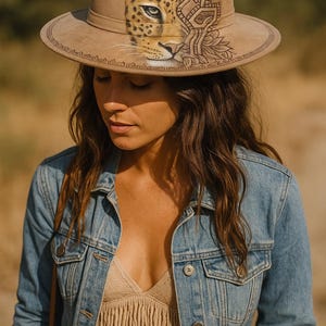 May include: A tan fedora hat with a detailed leopard design on the front. The hat features a wide brim and a decorative band around the crown. The hat is worn with a denim jacket and a fringed top.