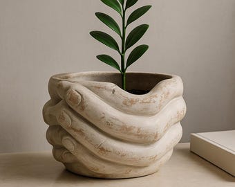 Pode incluir: Um vaso de cerâmica bege em forma de mãos, com uma planta verde no interior. O vaso tem um aspeto envelhecido e texturizado. A planta tem múltiplas folhas verdes. O vaso está sobre uma superfície clara.