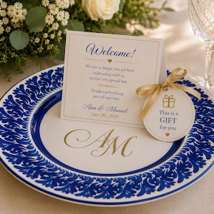 Puede incluir: Un plato de cerámica blanca con borde floral azul y monograma dorado AM, que incluye una tarjeta de bienvenida plegada con texto azul y dorado Welcome! Ana & Manuel June 20, 2025, y una etiqueta de regalo redonda atada con una cinta dorada.