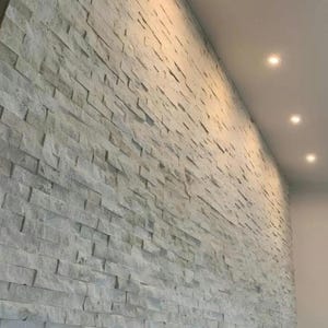 Puede incluir: Una pared texturizada hecha de ladrillos de piedra gris claro. Los ladrillos rectangulares están dispuestos en un patrón escalonado. Las luces empotradas en el techo iluminan la pared desde arriba, creando sombras y resaltando la textura.