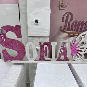 Puede incluir: Letras de purpurina rosa que deletrean "SOFIA" con un recorte de mariposa blanca y una base de madera blanca.