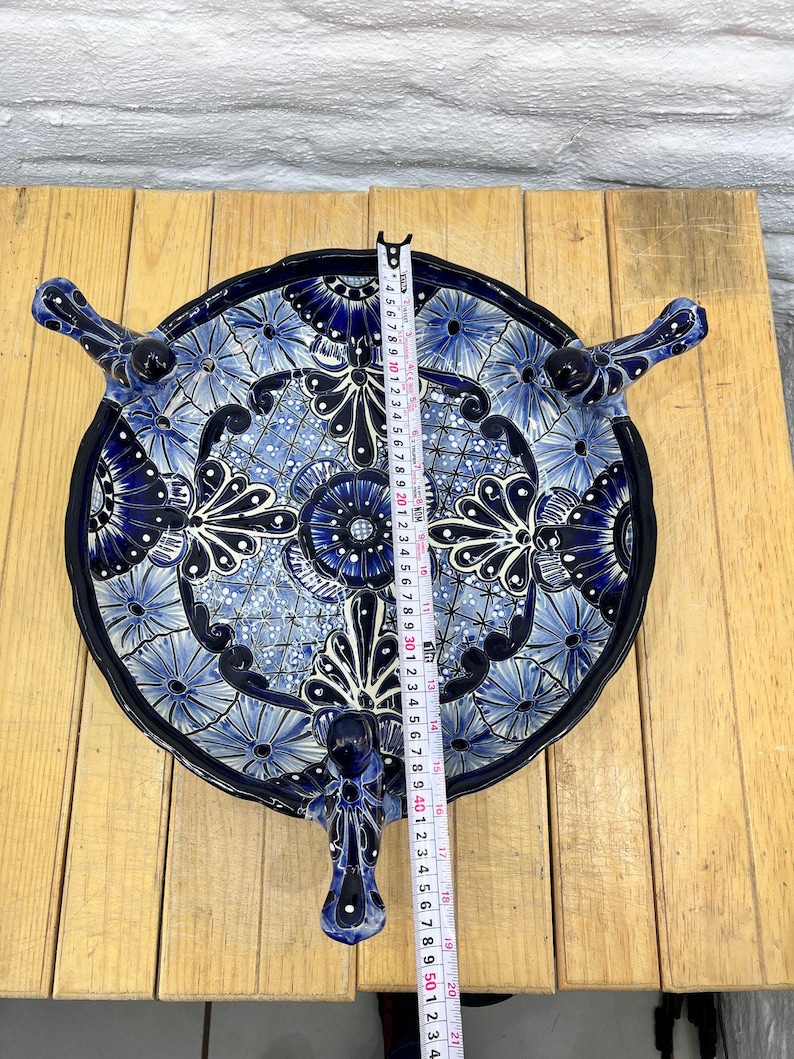 Pode incluir: Um prato de servir de cer&acirc;mica azul e branco com tr&ecirc;s pernas em forma de p&aacute;ssaros. O prato &eacute; decorado com um padr&atilde;o floral e tem um di&acirc;metro de aproximadamente 30 cm.