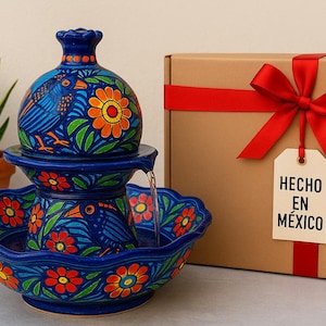 K&ouml;nnte beinhalten: Blaue Keramik-Brunnen mit Vogel- und Blumenmuster. Der Brunnen hat mehrere Ebenen und einen Wasserauslauf. Eine Geschenkbox mit rotem Band und einem Etikett mit der Aufschrift "HECHO EN M&Eacute;XICO" befindet sich daneben.
