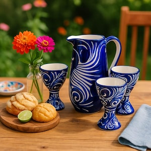Puede incluir: Una jarra de cerámica azul y blanca y copas a juego con un diseño de remolino. La jarra tiene un asa y un pico. Las copas tienen un tallo y un cuenco redondeado. El juego está sobre una mesa de madera con pan, lima y flores.