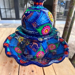K&ouml;nnte beinhalten: Ein lebendiger, handbemalter Keramikbrunnen mit blauem Sockel und aufwendigen Blumen- und Vogelmustern. Der Brunnen hat eine runde Oberseite und ein gewelltes Becken, wobei Wasser sanft von oben herabflie&szlig;t. Die Farben umfassen Blau, Rosa, Gelb und Orange.