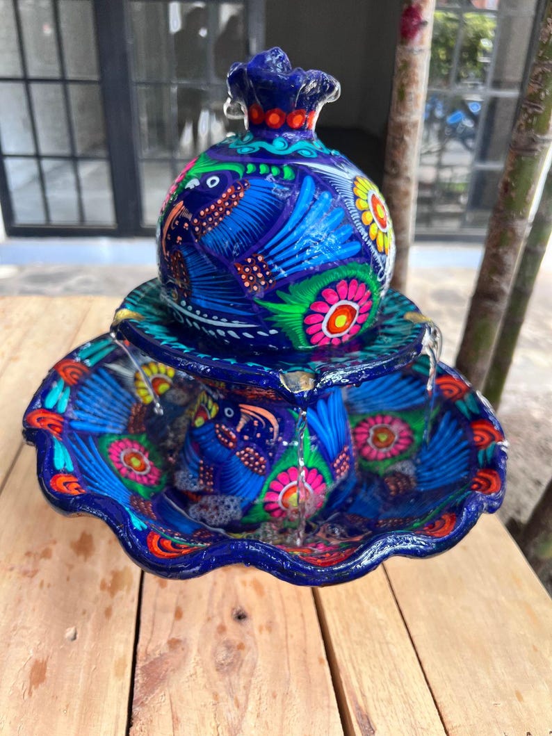 K&ouml;nnte beinhalten: Ein lebendiger, handbemalter Keramikbrunnen mit blauem Sockel und aufwendigen Blumen- und Vogelmustern. Der Brunnen hat eine runde Oberseite und ein gewelltes Becken, wobei Wasser sanft von oben herabflie&szlig;t. Die Farben umfassen Blau, Rosa, Gelb und Orange.