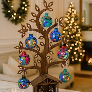 Puede incluir: Un árbol de Navidad de madera con adornos coloridos pintados a mano. El árbol tiene un pequeño nacimiento en la base. Los adornos son azules, verdes, rojos y amarillos. El árbol está sobre una superficie de mármol blanco.