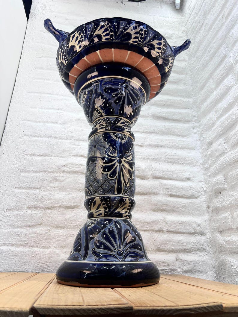 Pode incluir: Um bebedouro para p&aacute;ssaros de cer&acirc;mica azul e branco com um design floral decorativo. O bebedouro est&aacute; sobre um pedestal alto com uma base larga.