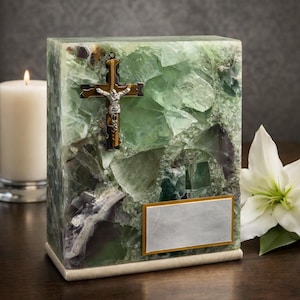 May include: A rectangular, green and purple stone urn with a gold and silver cross. The urn has a rectangular silver plate with a gold border. A white candle and flower are in the background.