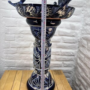 Pode incluir: Um bebedouro para p&aacute;ssaros de cer&acirc;mica azul e branco com dois p&aacute;ssaros empoleirados na borda. O bebedouro para p&aacute;ssaros est&aacute; em um suporte alto e decorativo com um padr&atilde;o floral azul e branco. O suporte mede 61 cm de altura.