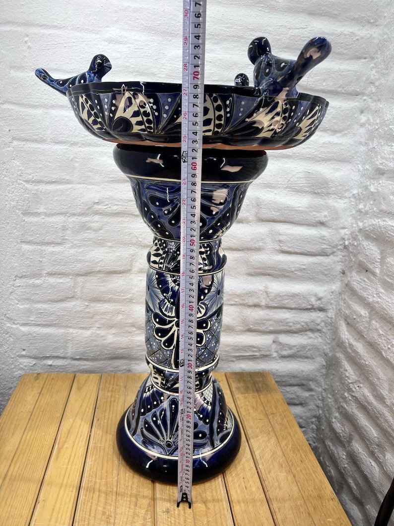 Pode incluir: Um bebedouro para p&aacute;ssaros de cer&acirc;mica azul e branco com dois p&aacute;ssaros empoleirados na borda. O bebedouro para p&aacute;ssaros est&aacute; em um suporte alto e decorativo com um padr&atilde;o floral azul e branco. O suporte mede 61 cm de altura.