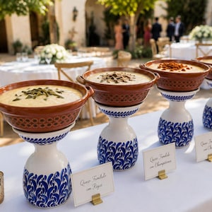 Puede incluir: Una mesa de buffet en un evento al aire libre presenta varios cuencos de comida. Los cuencos son marrones y se asientan sobre pedestales decorativos blancos y azules. Cada cuenco contiene un adorno diferente. Pequeños letreros identifican los platos.