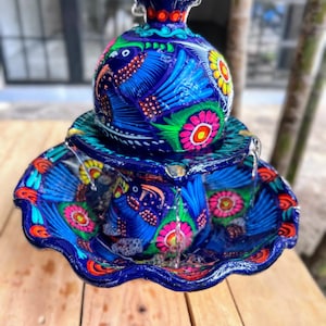 K&ouml;nnte beinhalten: Handbemalter Keramikbrunnen mit blauem Sockel und Schale. Der Brunnen zeigt farbenfrohe Vogel- und Blumendesigns in Pink, Gelb und Gr&uuml;n. Die Oberseite hat einen dekorativen Aufsatz.