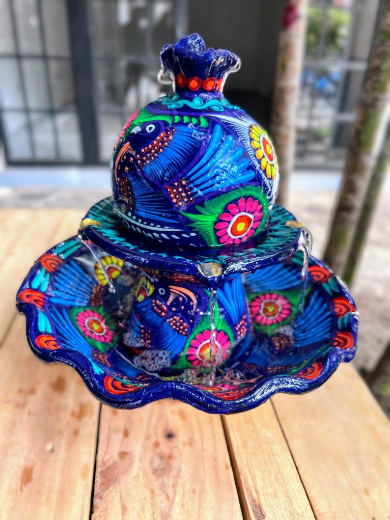 K&ouml;nnte beinhalten: Handbemalter Keramikbrunnen mit blauem Sockel und Schale. Der Brunnen zeigt farbenfrohe Vogel- und Blumendesigns in Pink, Gelb und Gr&uuml;n. Die Oberseite hat einen dekorativen Aufsatz.