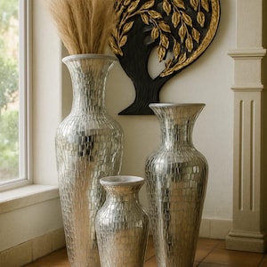 May include: Three silver mosaic-textured vases of varying heights, with a tall vase holding dried ornamental grass. A black and gold tree-shaped wall hanging is in the background. The vases are decorative home decor.