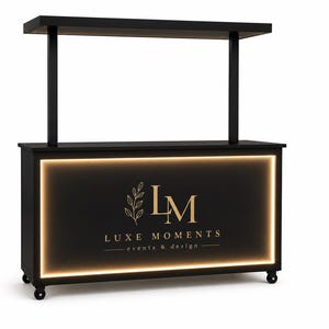 May include: A black rectangular event bar with a top shelf supported by two vertical posts. The front panel features a glowing rectangular light border and the gold text "LM LUXE MOMENTS events & design" with a leaf graphic. It stands on small wheels.