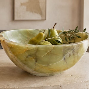 May include: A large, round, translucent green onyx stone bowl with natural veining and polished edges. Inside the bowl are three fresh green pears and a small sprig of green leaves, showcasing the smooth, marbled texture of the stone material.