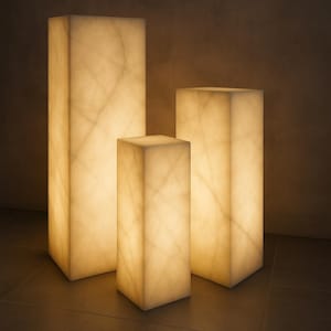 Puede incluir: Tres pedestales rectangulares iluminados con un interior cálido y brillante. Los pedestales varían en altura, el más alto a la izquierda y el más bajo en el centro. Las superficies tienen un patrón veteado, lo que sugiere un material de piedra natural.