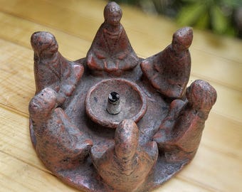 May include: A terracotta incense burner shaped like a circle with eight figures sitting around the edge. The figures are all facing inward towards a central incense holder. The burner is a reddish-brown color and has a rustic, hand-crafted look.