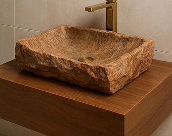 May include: Hand-carved Mexican marble rectangular sink with a rough, natural texture. The sink is a warm, earthy tone and sits on a dark wood floating vanity. A gold-toned faucet is mounted on the wall. The sink measures 19 x 15 inches.