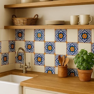 Puede incluir: Interior de cocina con fregadero blanco, encimeras de madera y armarios color crema. El salpicadero presenta baldosas estampadas con diseños florales azules y naranjas. Estantes de madera exhiben cuencos, tazas y una cesta. También hay una planta en maceta y utensilios de madera.