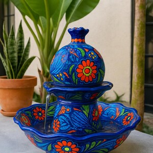 K&ouml;nnte beinhalten: Blauer Keramikbrunnen mit floralem Design. Der Brunnen hat einen runden Aufsatz und eine gewellte Schale, verziert mit orangefarbenen Blumen und gr&uuml;nen Bl&auml;ttern. Wasser flie&szlig;t von oben und schafft ein dekoratives Element f&uuml;r den Au&szlig;enbereich.