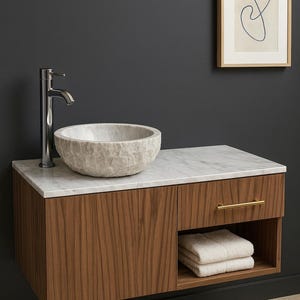 Puede incluir: Mueble de baño moderno con encimera de mármol y lavabo de piedra texturizada. El mueble tiene un acabado de madera oscura con un tirador dorado. Un grifo cromado y una obra de arte abstracta enmarcada completan el diseño.
