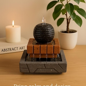 Könnte beinhalten: Indoor-Brunnen mit einer strukturierten, schwarzen Kugel auf einem mehrstufigen, quadratischen Sockel. Der Sockel besteht aus braunem und grauem Stein. Eine brennende Kerze und eine Topfpflanze befinden sich im Hintergrund. Der Text lautet: Bringen Sie Ruhe und Harmonie in Ihr modernes Zuhause.