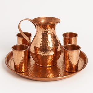 Puede incluir: Una jarra de cobre martillado con asa, rodeada de cuatro vasos de cobre a juego, todo sobre una bandeja redonda de cobre. El cobre tiene un tono marrón rojizo cálido y una superficie texturizada y con hoyuelos.