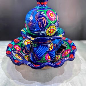 K&ouml;nnte beinhalten: Handbemaltes Keramik-Set mit einer blauen Vase mit Deckel, einer Tasse und einem Teller. Das Set ist mit bunten Blumen- und Vogelmotiven in Rosa, Gelb, Gr&uuml;n und Rot verziert. Mit gewelltem Rand.