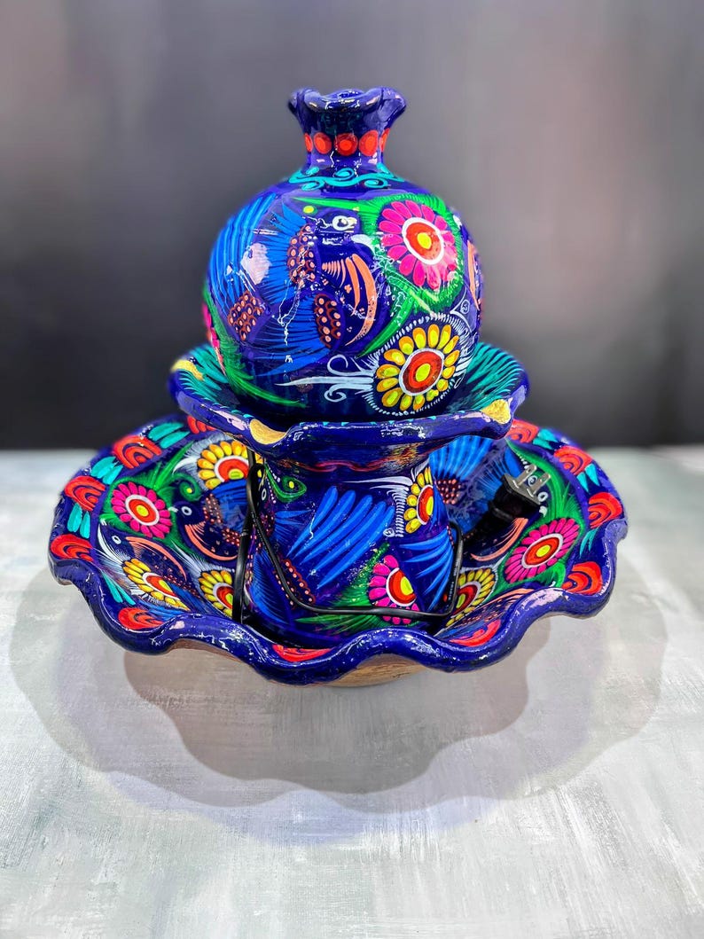 K&ouml;nnte beinhalten: Handbemaltes Keramik-Set mit einer blauen Vase mit Deckel, einer Tasse und einem Teller. Das Set ist mit bunten Blumen- und Vogelmotiven in Rosa, Gelb, Gr&uuml;n und Rot verziert. Mit gewelltem Rand.
