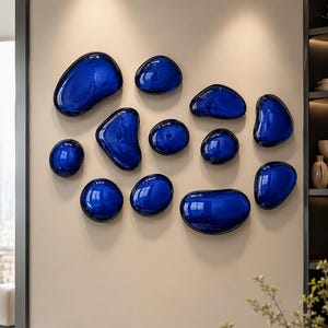 Puede incluir: Una colección de once decoraciones de pared con forma de guijarro, de color azul intenso y brillante, dispuestas en grupo. Cada pieza presenta una forma orgánica, lisa y redondeada con una superficie reflectante que capta la luz.