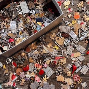 May include: A selection of metal charms in a wooden box and on a wooden surface. The charms feature diverse shapes and designs, including hearts, stars, and miniature objects. The text "THIS IS A POSITIVE SIGN" is visible on a white charm.