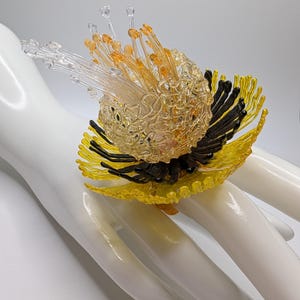 May include: A large, intricate ring featuring a central cluster of clear and amber glass filaments. The base is composed of radiating yellow glass petals interspersed with dark, black glass stamens, creating a multi-layered, textured floral design.