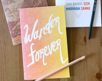 May include: Two small notebooks with colorful covers. One notebook has a mountain scene and the text "wild & free". The other notebook has a gradient background and the text "Wander forever". A third card has a list of greetings in different languages.  Two pencils are on the table.