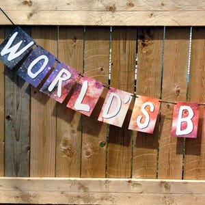 Puede incluir: Una guirnalda con las palabras "WORLD'S BEST" en letras may&uacute;sculas blancas sobre un fondo de galaxia colorido. La guirnalda est&aacute; colgada frente a una valla de madera.