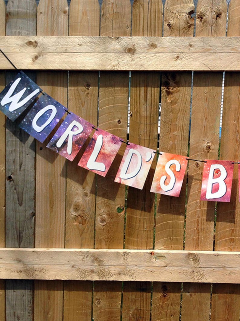 Puede incluir: Una guirnalda con las palabras "WORLD'S BEST" en letras may&uacute;sculas blancas sobre un fondo de galaxia colorido. La guirnalda est&aacute; colgada frente a una valla de madera.