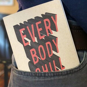 May include: A brown notebook with the text "EVERYBODY CHILL" printed in black and red letters. The notebook is in a back pocket of a pair of dark gray pants.