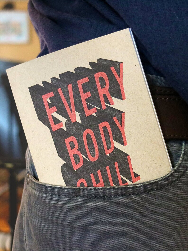 May include: A brown notebook with the text "EVERYBODY CHILL" printed in black and red letters. The notebook is in a back pocket of a pair of dark gray pants.