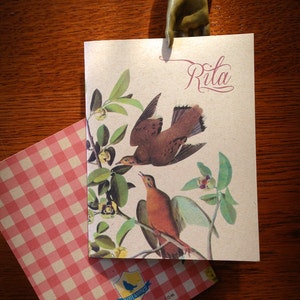 May include: A light brown card with a brown and white bird illustration and green leaves. The card has the name "Rita" written in cursive on the top right corner. The card is on a wooden surface with a metal hand holding it in place.