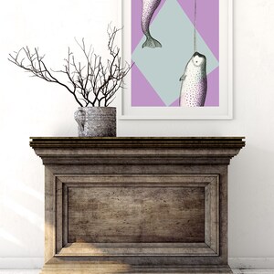 May include: A framed art print featuring a narwhal illustration with a long tusk, set against a geometric background of purple and light blue. The artwork is displayed on a wooden cabinet, with a decorative branch arrangement in a metal pot.