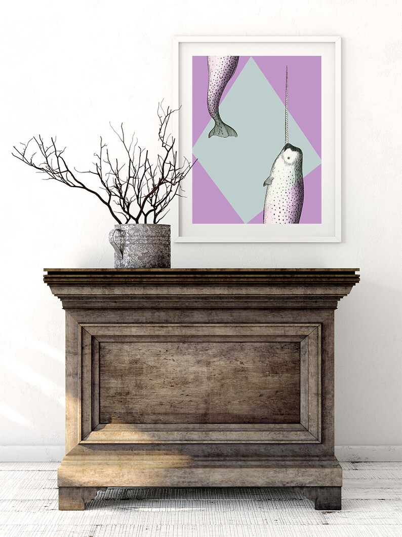 May include: A framed art print featuring a narwhal illustration with a long tusk, set against a geometric background of purple and light blue. The artwork is displayed on a wooden cabinet, with a decorative branch arrangement in a metal pot.
