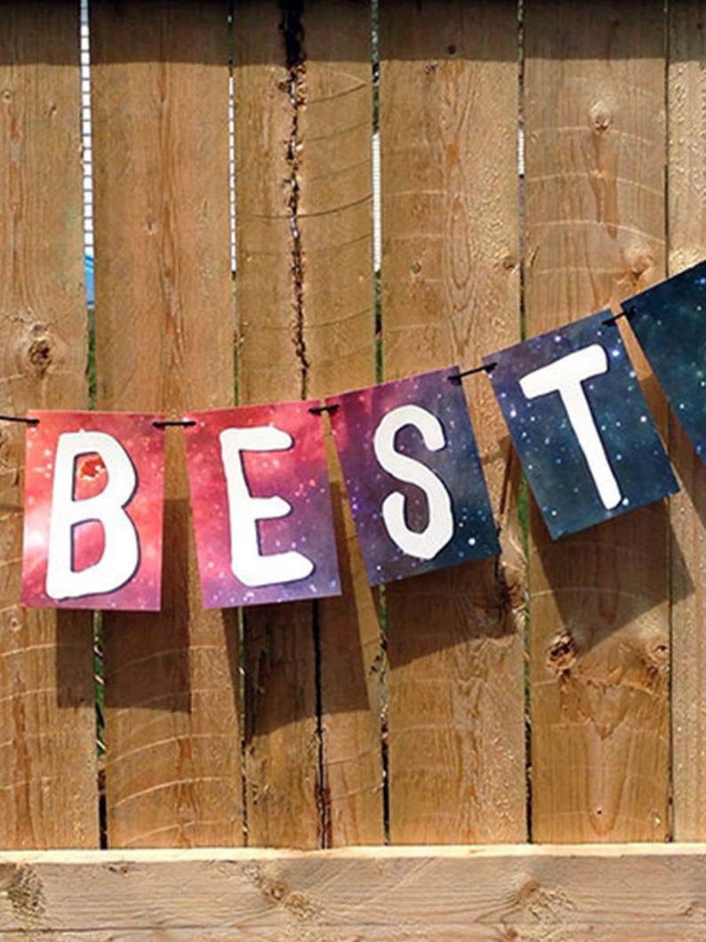 Puede incluir: Una guirnalda decorativa con la palabra "BEST" escrita en letras blancas sobre un fondo de galaxia colorida. La guirnalda est&aacute; colgada frente a una valla de madera, creando un ambiente festivo.