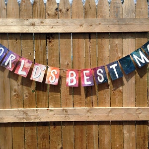 Puede incluir: Una pancarta colgada sobre una valla de madera, con las palabras "WORLD'S BEST MOM" escritas en banderas rectangulares individuales y coloridas. Las banderas tienen un fondo similar a una galaxia con letras may&uacute;sculas blancas. La pancarta est&aacute; suspendida por una fina cuerda negra.