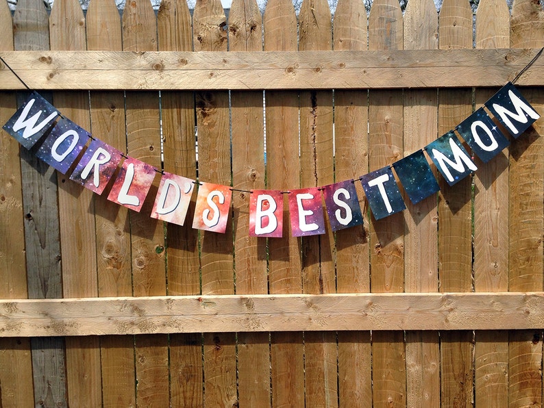 Puede incluir: Una pancarta colgada sobre una valla de madera, con las palabras "WORLD'S BEST MOM" escritas en banderas rectangulares individuales y coloridas. Las banderas tienen un fondo similar a una galaxia con letras may&uacute;sculas blancas. La pancarta est&aacute; suspendida por una fina cuerda negra.